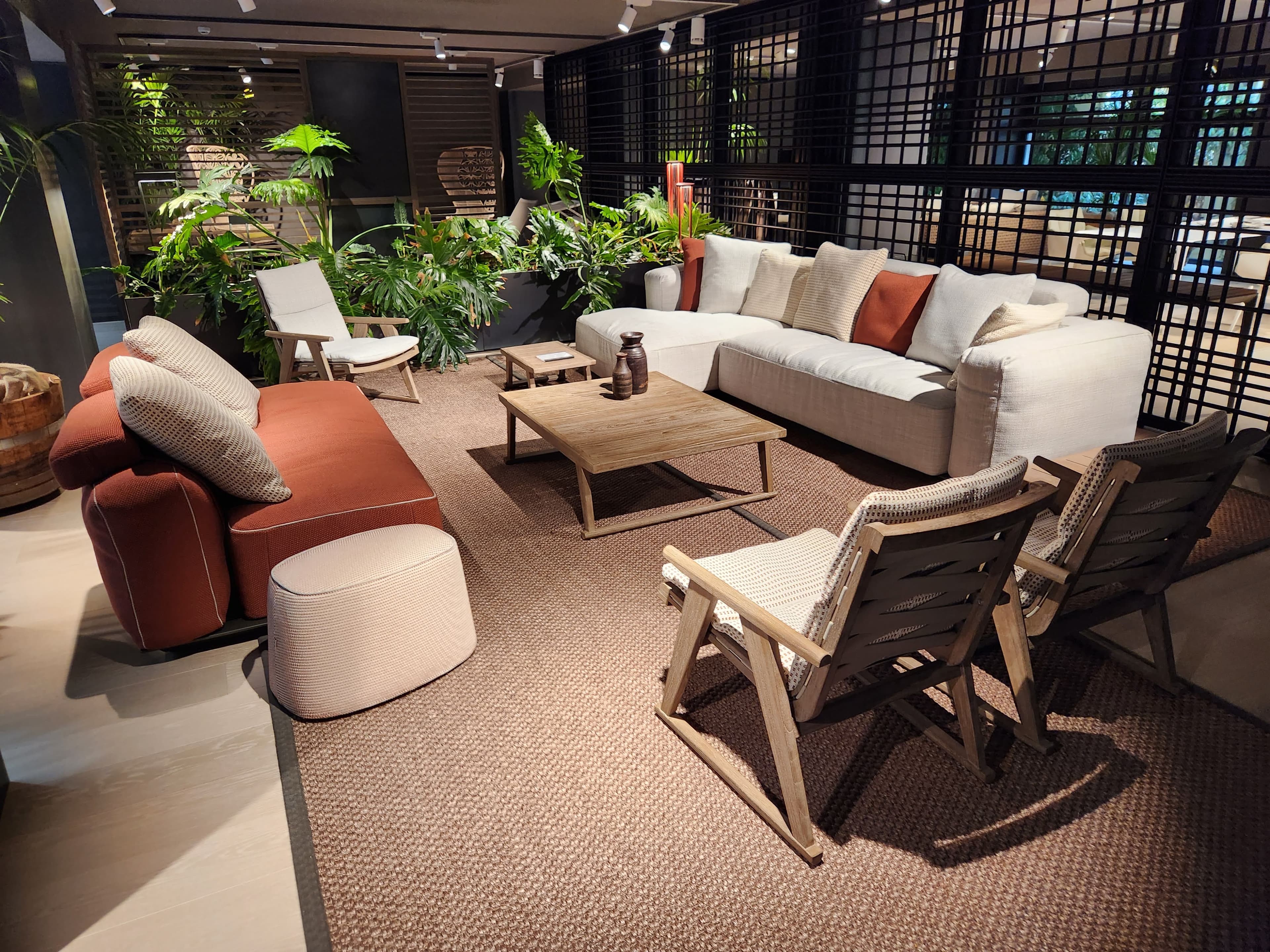 A sophisticated showroom interior showcasing a living room arrangement with a large L-shaped sofa, a smaller terracotta sofa, wooden coffee tables, and lounge chairs, all set on a natural fiber rug amidst lush green plants.