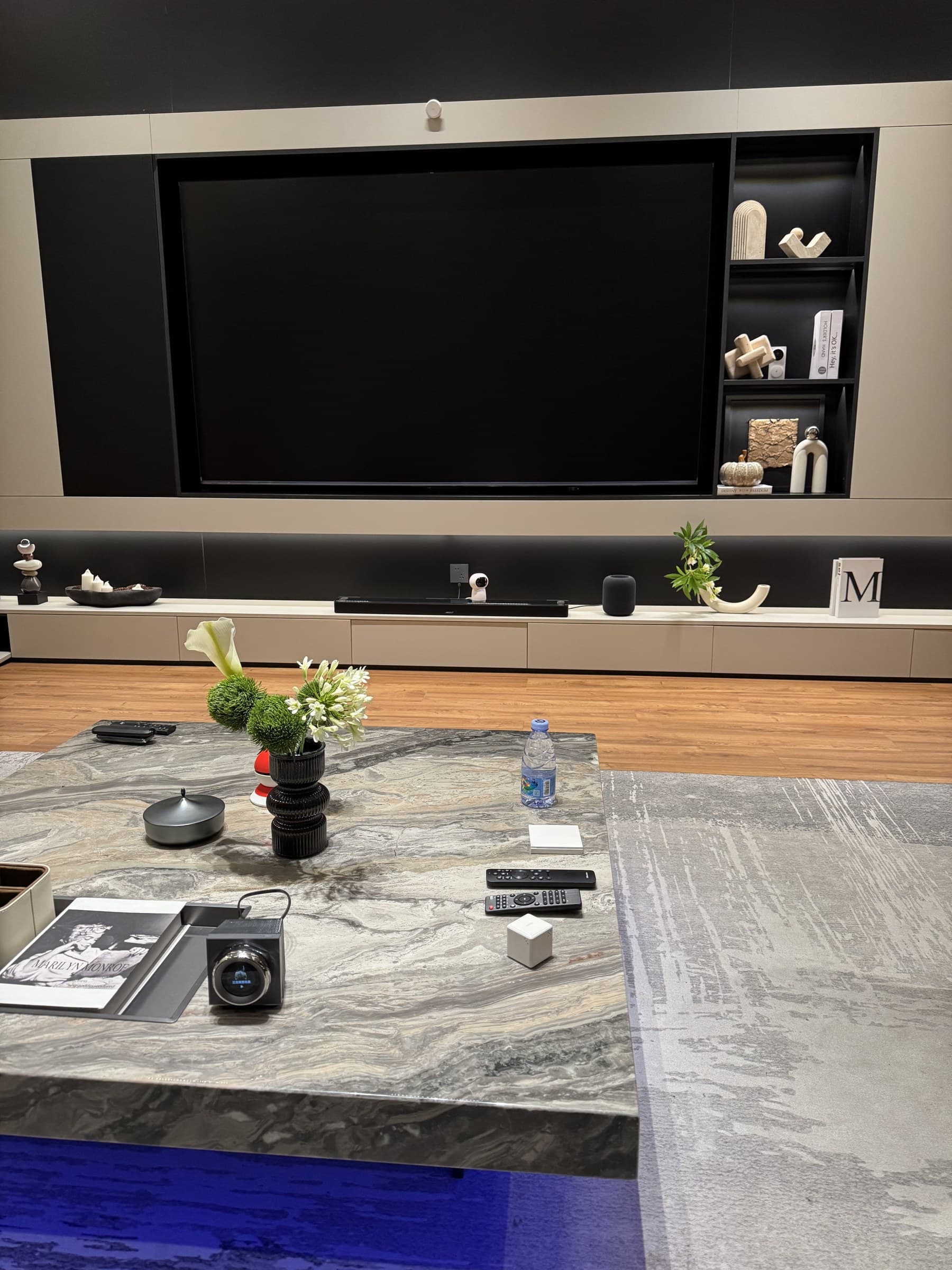 A modern living room with a large TV, soundbar, and decorative shelves. A marble coffee table in the foreground holds a vase of flowers, remotes, a water bottle, and smart home devices.