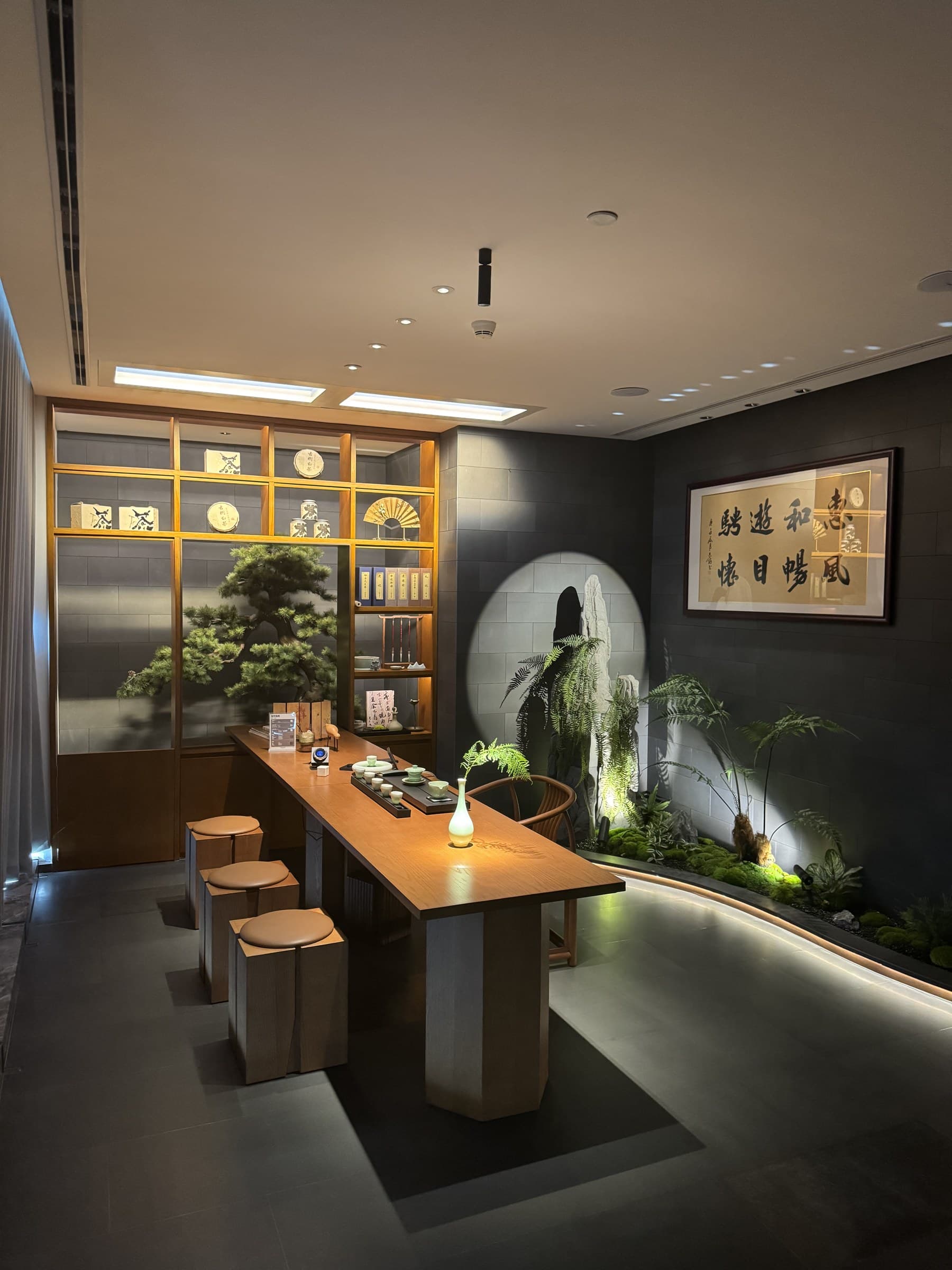 A modern room with a long wooden table, stools, a display shelf, and an indoor garden with illuminated plants and rocks.