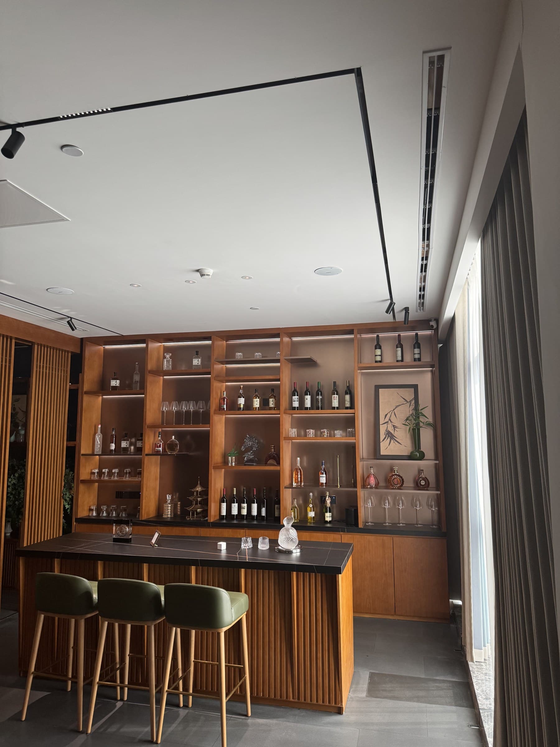 A stylish bar area with a large wooden shelving unit displaying bottles and glasses, a dark bar counter, and three green bar stools. The ceiling has modern lighting fixtures and a smoke detector.