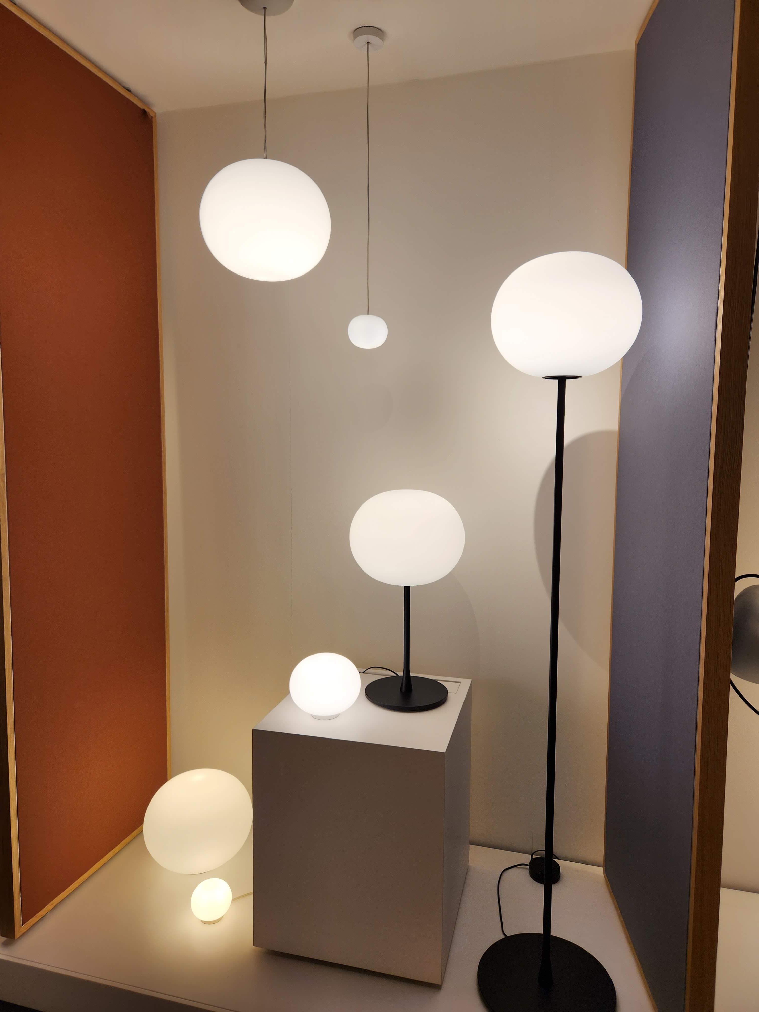A modern showroom interior showcasing a collection of spherical white lamps, including pendant lights, a floor lamp, and various table lamps, displayed against a warm beige wall and framed panels in terracotta and muted purple.